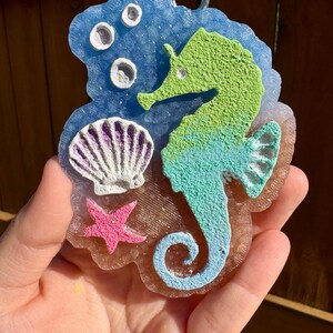 May include: A resin seahorse ornament with a blue and green color scheme. The ornament features a seahorse, a seashell, a starfish, and bubbles. The seahorse is green and blue, the seashell is white and purple, and the starfish is pink.