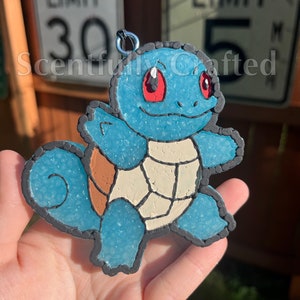 以下が含まれることがあります： 赤い目をした青と茶色のゼニガメのポケモンチャーム。チャームはポリマークレイ製で、表面にテクスチャがあります。チャームは銀色のリングにぶら下がっています。