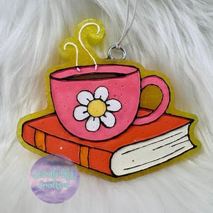 May include: A resin hanging ornament featuring a pink mug with a white flower design resting on an orange book. The mug has steam rising from it, and the entire piece is outlined in a yellow border with a silver metal loop for hanging.