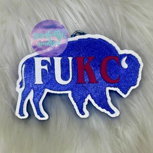 May include: A blue glittery buffalo-shaped keychain with a white border. The text FUKC is printed across the center in white and red letters. A circular logo with the text Scentfully Crafted is visible in the upper left corner.