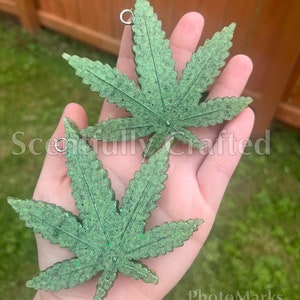 May include: Two green, glitter-infused cannabis leaf pendants with silver-colored jump rings. The pendants are held in a hand against a blurred outdoor background. The text "Scoutfully Crafted" is visible in the image.