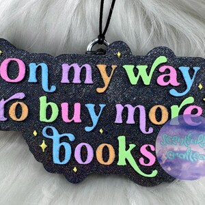 May include: A black glittery hanging ornament with a cloud-like shape featuring the colorful text On my way to buy more books in pink, blue, orange, and green. Small yellow star accents are scattered across the surface with a black string loop.