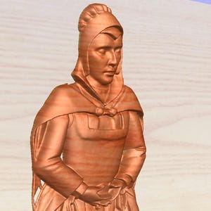 May include: A wooden sculpture of a woman wearing a hooded cloak and dress. The figure is carved from wood, with visible grain and a warm, reddish-brown color. The woman's hands are clasped in front of her.