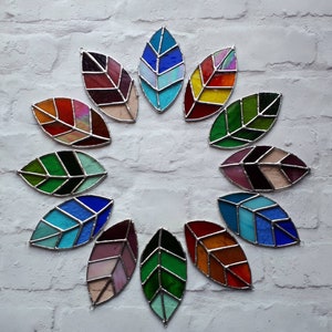 Op de afbeelding: Een set van 12 gebrandschilderde bladeren in verschillende kleuren, waaronder rood, blauw, groen en geel. De bladeren zijn in een cirkel gerangschikt, met de punten van de bladeren naar buiten gericht.