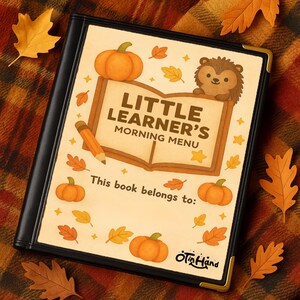 May include: A black binder with gold corner protectors, featuring a fall-themed cover. The cover reads "Little Learner's Morning Menu" with illustrations of pumpkins, leaves, a pencil, and a hedgehog. The text "This book belongs to:" is also visible.