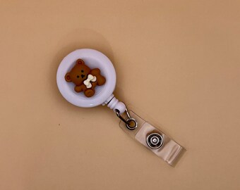May include: White retractable badge holder with a brown teddy bear charm holding a bone. The charm is attached to a white circle with a clear plastic strap.