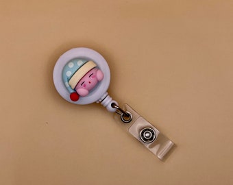 May include: A white retractable badge reel with a sleeping pink character wearing a blue polka-dot hat. The character has a beige brim on the hat and a red ball on the side. The reel has a clear plastic clip.