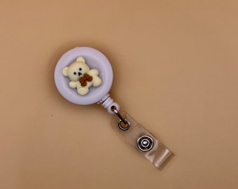 May include: A white retractable badge reel with a cute teddy bear design. The teddy bear has a brown bow and is centered on the reel. The reel has a clear plastic strap and a metal clip.