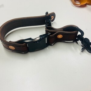 May include: A dark brown leather strap featuring black stitching, secured with a black plastic side-release buckle. The strap includes copper-colored metal rivets and a black metal swivel lobster clasp attached to one end for secure fastening.