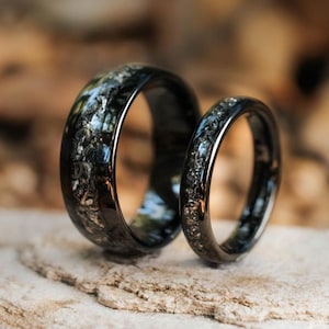May include: Two black rings with a wide and a narrow band, both featuring a textured inlay of dark, metallic, and shimmering particles embedded within the centre of the polished black metal surface of each circular band.