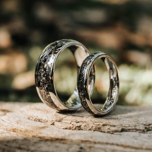 Peut inclure: Deux bagues argentées avec une finition polie et une incrustation centrale composée d'un matériau sombre, texturé et parsemé de reflets métalliques. La bague la plus large est placée derrière la plus fine, toutes deux présentant un design cohérent avec des particules sombres et irrégulières incrustées au centre de l'anneau.