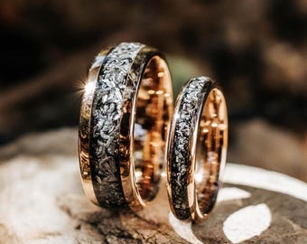 May include: Two rings with a rose gold finish and a central inlay featuring a textured, metallic silver and dark gray crystalline pattern. The bands have a polished, reflective surface and a rounded profile, showcasing a distinct contrast between the warm metal edges and the intricate, sparkling center inlay.