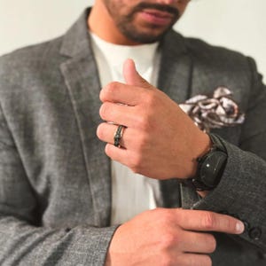 May include: A black and gold ring with a textured band. The ring is worn on the left ring finger of a person wearing a gray blazer.