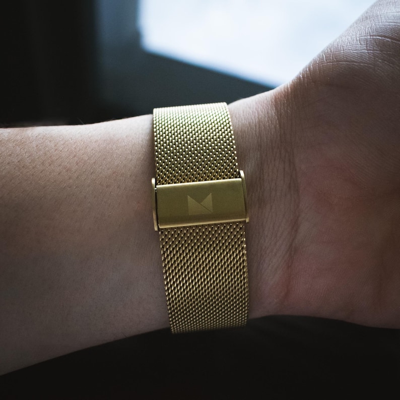 May include: A gold metal watch strap with a mesh design. The strap has a small, rectangular buckle with a logo on it.