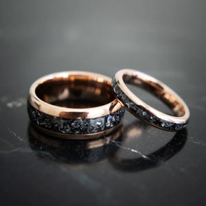 May include: Two rose gold wedding bands with a black inlay. The bands are made of tungsten carbide and have a brushed finish.