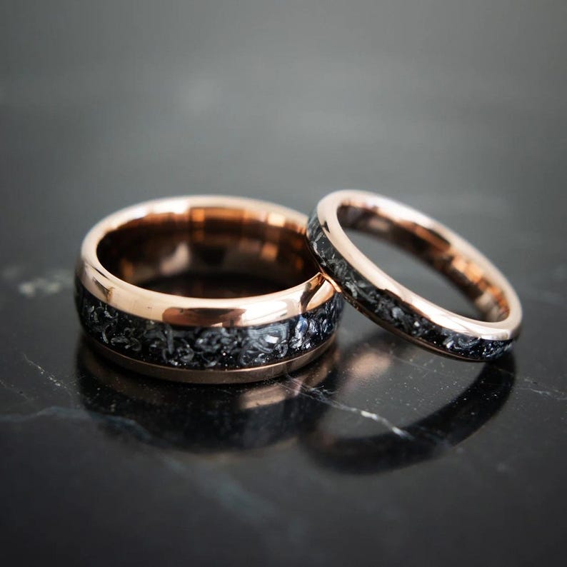 May include: Two rose gold wedding bands with a black inlay. The bands are made of tungsten carbide and have a brushed finish.
