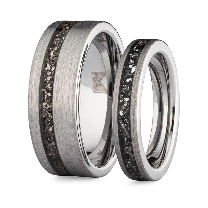 May include: A pair of silver wedding bands with a black meteorite inlay. The band on the left has a wider band and a single inlay, while the band on the right has a thinner band and a single inlay.