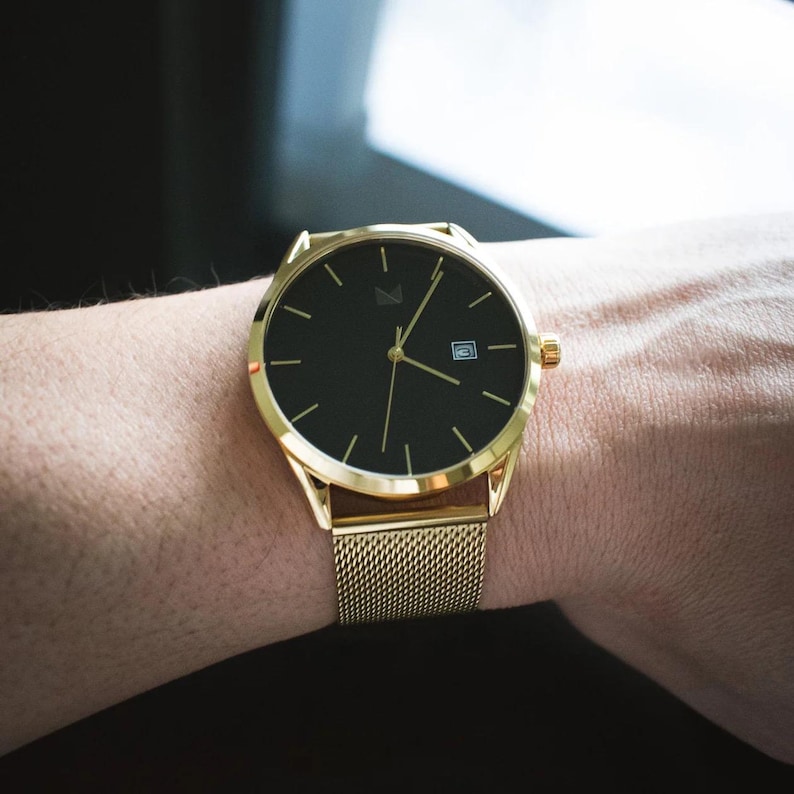 May include: A gold-tone watch with a black face and a mesh metal strap. The watch has a date window at the 3 o'clock position.