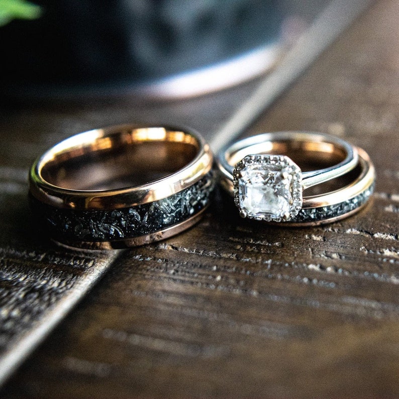 May include: A pair of wedding bands, one with a black and gray speckled finish and a rose gold band, and the other with a diamond ring with a halo setting.