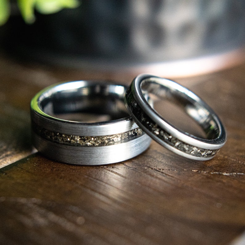 May include: Two silver wedding bands with a black inlay. The bands are on a wooden surface.