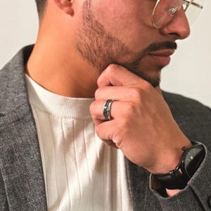 K&ouml;nnte beinhalten: Ein silberner Ring mit einem schwarzen Band und einem silbernen Band mit einem strukturierten Design. Der Ring wird am Ringfinger der linken Hand einer Person getragen.
