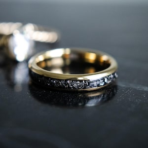 May include: A gold wedding band with a black meteorite inlay. The band is polished and has a smooth finish.
