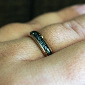 May include: A black and gold ring with a textured black band. The ring is worn on a person's finger.