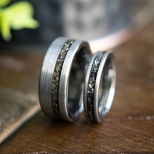 May include: Two silver wedding bands with a textured, dark gray inlay. The bands are stacked on top of each other.