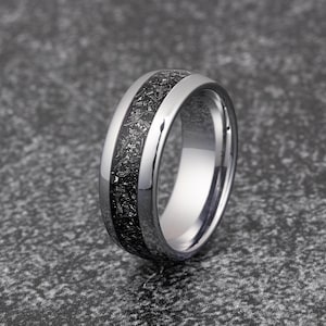 K&ouml;nnte beinhalten: Ein silberner Wolframcarbid-Ring mit einer schwarzen Einlage. Die Einlage hat ein gesprenkeltes Muster.