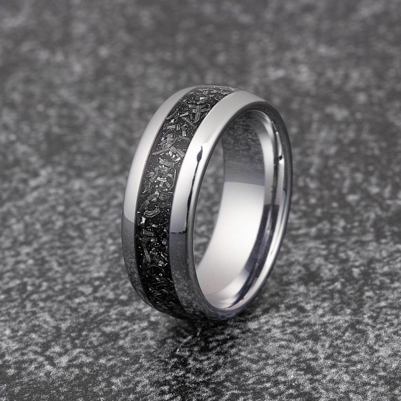K&ouml;nnte beinhalten: Ein silberner Wolframcarbid-Ring mit einer schwarzen Einlage. Die Einlage hat ein gesprenkeltes Muster.