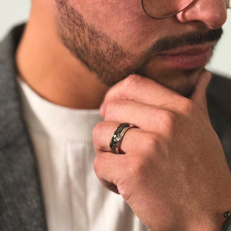 May include: A black and gold ring with a textured band. The ring is worn on the ring finger of a person's hand.