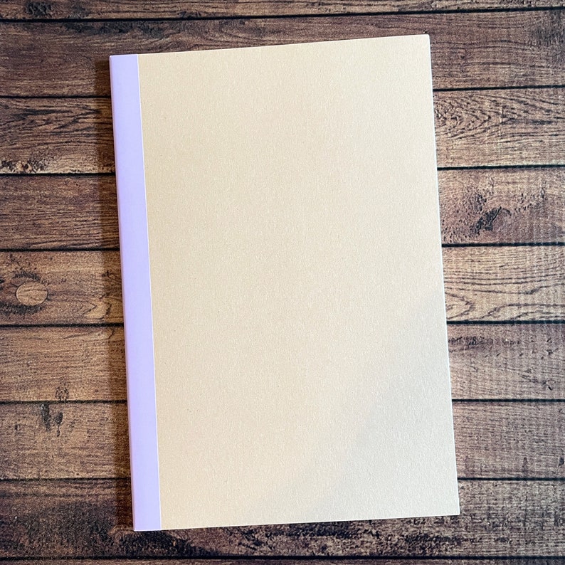 May include: A cream-colored notebook with a lavender spine. The notebook is rectangular and has a textured surface. The background is a wooden surface.