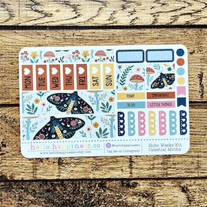 May include: A sticker sheet with a celestial moth theme. The sheet includes stickers for each day of the week, plus decorative elements like mushrooms and flowers. The color palette includes blues, yellows, and pinks. The text "hello.happiness.co" is visible.