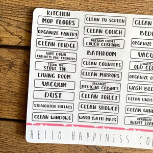 May include: A sheet of white cleaning labels with black text. Labels include "Kitchen", "Mop Floors", "Clean Fridge", "Vacuum", and "Clean Windows". The bottom of the sheet has the text "Hello Happiness Co" in a handwritten font.