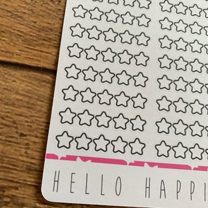 Puede incluir: Una hoja de papel adhesivo blanco con una franja rosa en la parte inferior. El papel est&aacute; lleno de pegatinas de estrellas de contorno negro. El texto "HELLO HAPPY" est&aacute; impreso en la parte inferior de la hoja.