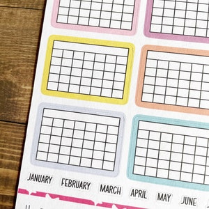 Pu&ograve; includere: Sei adesivi colorati a griglia con i mesi dell'anno etichettati sotto: gennaio, febbraio, marzo, aprile, maggio e giugno. Gli adesivi sono su una superficie di legno. Il testo "HELLO HAPPINESS" &egrave; stampato sotto gli adesivi.