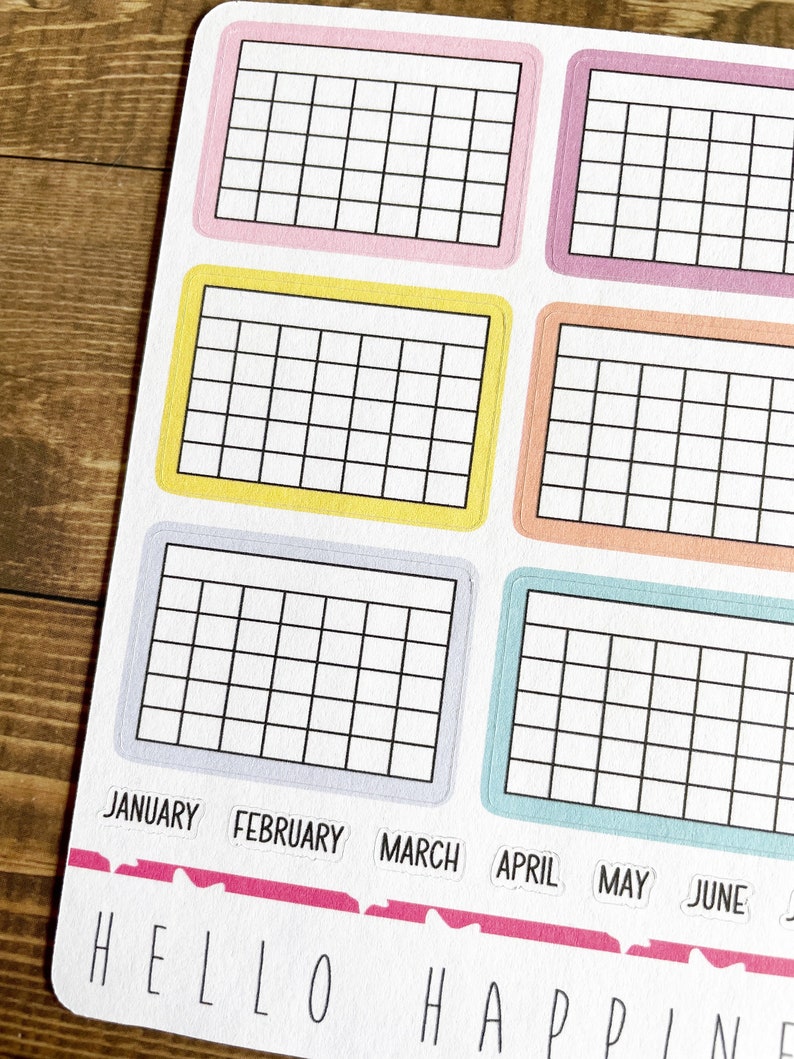 Pu&ograve; includere: Sei adesivi colorati a griglia con i mesi dell'anno etichettati sotto: gennaio, febbraio, marzo, aprile, maggio e giugno. Gli adesivi sono su una superficie di legno. Il testo "HELLO HAPPINESS" &egrave; stampato sotto gli adesivi.