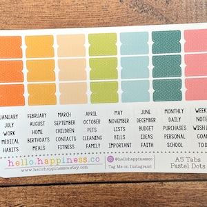 Puede incluir: Una hoja de pestañas de planificador A5 de color pastel con puntos blancos. Las pestañas están etiquetadas con los meses del año, los días de la semana y varias categorías como "trabajo", "médico", "hogar", "facturas", "presupuesto", "escuela" y "metas".