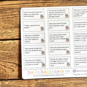 May include: A sheet of stickers with mindfulness prompts in black text on white rectangles. The stickers feature questions like "List five activities that make you feel calm" and include a hand with a peace sign. The website hello.happiness.co is visible.