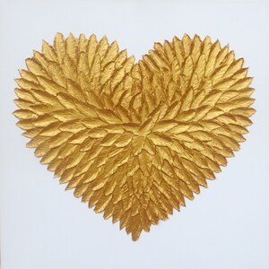 May include: A heart-shaped wall art piece composed of numerous individual gold-colored feather-like elements arranged in a dense, layered pattern. The metallic gold texture creates a three-dimensional effect against a plain white background.