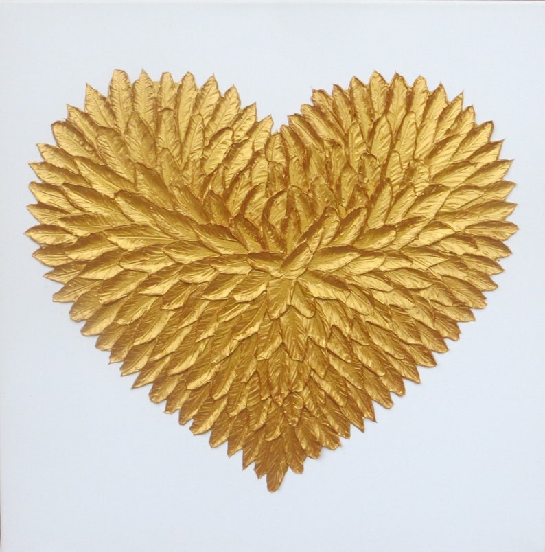 May include: A heart-shaped wall art piece composed of numerous individual gold-colored feather-like elements arranged in a dense, layered pattern. The metallic gold texture creates a three-dimensional effect against a plain white background.