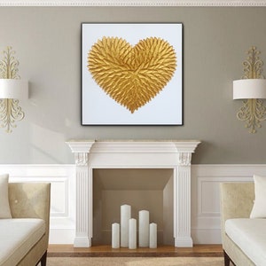 May include: A square wall art piece featuring a textured, three-dimensional golden heart shape composed of numerous small, leaf-like elements. The heart is centered on a plain white background and enclosed within a thin, black frame.