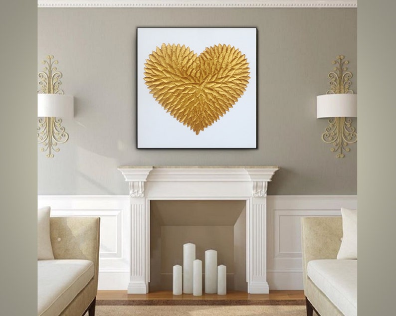 May include: A square wall art piece featuring a textured, three-dimensional golden heart shape composed of numerous small, leaf-like elements. The heart is centered on a plain white background and enclosed within a thin, black frame.