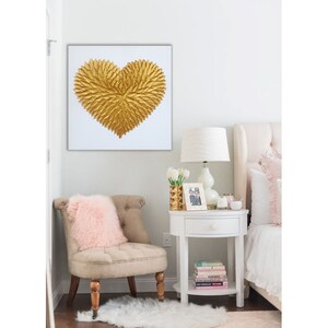 May include: A square wall art piece featuring a textured, three-dimensional gold heart shape composed of numerous small, leaf-like or feather-like elements arranged in a radiating pattern against a plain, solid white background.