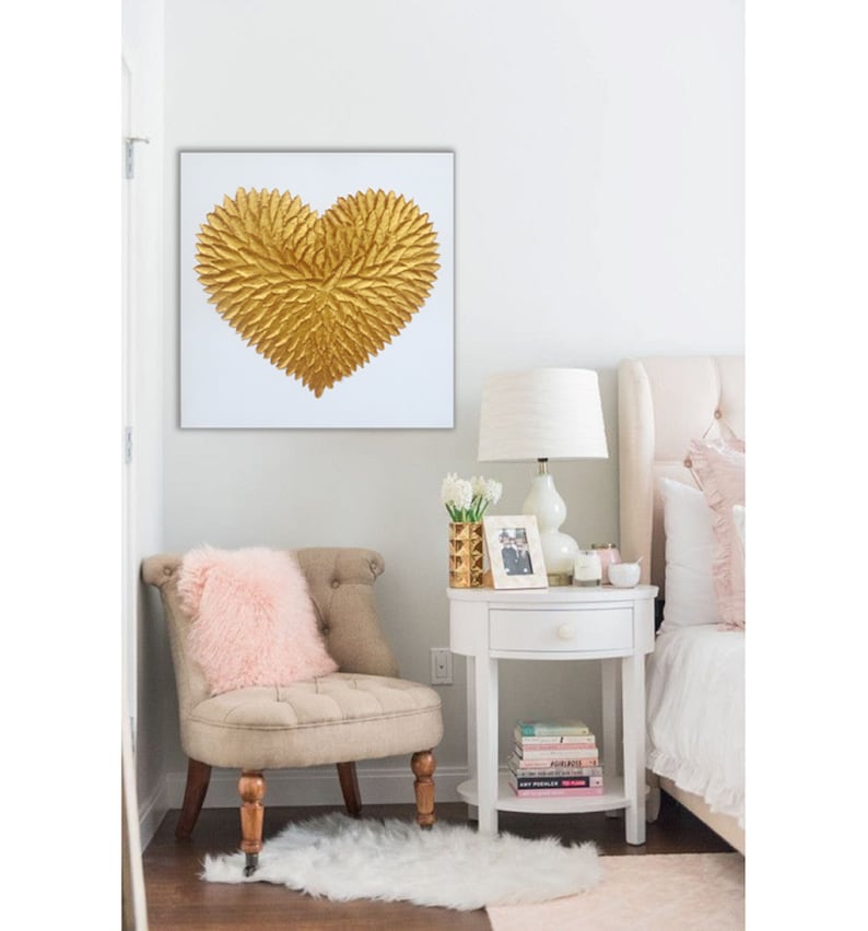 May include: A square wall art piece featuring a textured, three-dimensional gold heart shape composed of numerous small, leaf-like or feather-like elements arranged in a radiating pattern against a plain, solid white background.