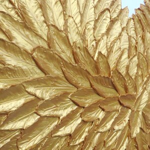 May include: A decorative wall art piece featuring a dense arrangement of overlapping, textured gold-painted feathers. The feathers are layered to create a wing-like shape with a metallic finish, showing intricate vein details on each individual element.