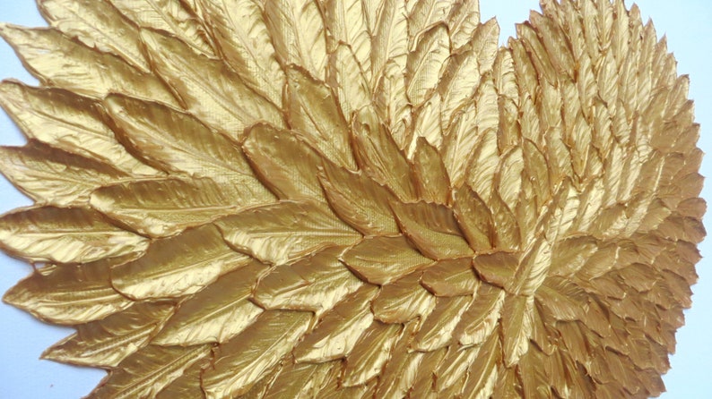 May include: A decorative wall art piece featuring a dense arrangement of overlapping, textured gold-painted feathers. The feathers are layered to create a wing-like shape with a metallic finish, showing intricate vein details on each individual element.