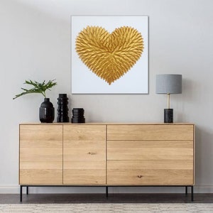 May include: A square wall art piece featuring a textured, three-dimensional gold heart shape composed of numerous leaf-like elements centered on a plain white background. The artwork is displayed above a light wood sideboard with black metal legs.