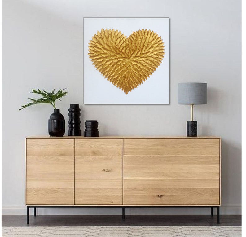 May include: A square wall art piece featuring a textured, three-dimensional gold heart shape composed of numerous leaf-like elements centered on a plain white background. The artwork is displayed above a light wood sideboard with black metal legs.