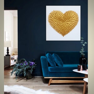 May include: A square wall art piece featuring a textured, three-dimensional gold heart composed of layered leaf-like shapes centered on a plain white background. The artwork is displayed on a dark navy blue wall above a deep blue velvet sofa.